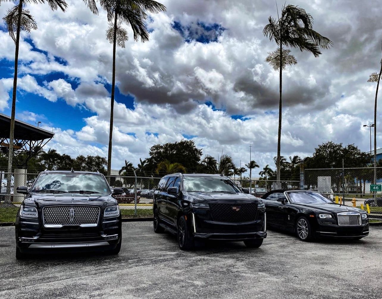 Three black cars parked in a parking lot under some clouds.